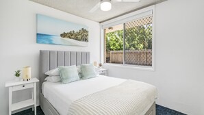Een slaapkamer met een bed, nachtkastje, plafondventilator, raam met uitzicht op groen, en een schilderij aan de muur.