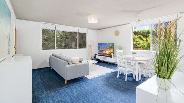Een moderne woonkamer met een grijze bank, een witte salontafel en een eetgedeelte met een witte tafel en stoelen. Er is een groot raam met uitzicht op groen.