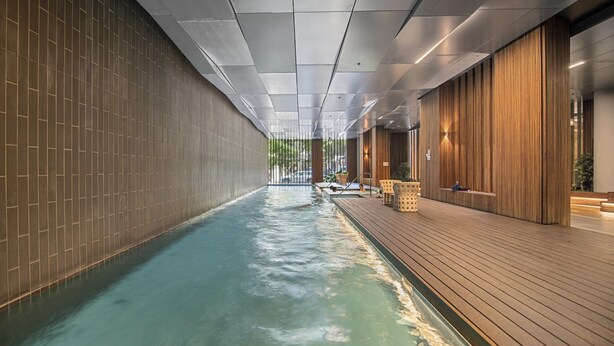 Una piscina interior moderna con una terraza de madera, paredes de azulejos e iluminación empotrada en el techo.