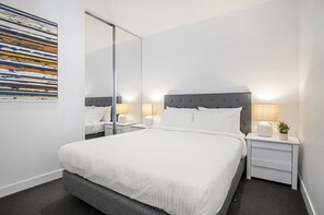 Une chambre d’hôtel moderne avec un grand lit, des tables de chevet avec des lampes, un miroir et un tableau abstrait aux couleurs vives accroché au mur.