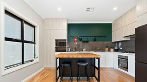 A modern kitchen with a central island, bar stools, a built-in oven, and a window with plants. - Bright 2-bed Townhouse With Balcony & Garage (Adelaide)