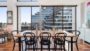 Un comedor moderno con mesa de madera y sillas negras, ventanas amplias con vistas a la ciudad y una decoración minimalista con cuadros enmarcados y una mesita auxiliar.