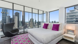 Habitación de hotel en un rascacielos con una cama grande, una silla moderna y vistas a la ciudad a través de ventanales del piso al techo.
