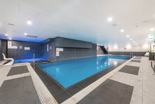 An indoor swimming pool with a blue water feature, a diving board, and a seating area with a chair and a plant. - Incredible City Views in Convenient CBD Apartment (Melbourne)
