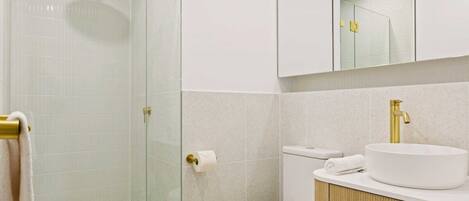 A modern bathroom with a glass shower enclosure, a white toilet, and a vanity with a gold faucet and sink.