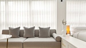 A modern hotel room with a neutral color scheme, featuring a sofa with three cushions, a bedside table, a lamp, and a white dresser with gold handles. - City Centre Studio by Hyde Park (Sydney)