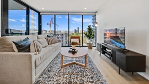 A modern living room with a sofa, coffee table, TV, and a large window with a city view. - Chic 1-bed With Sophisticated Decor, (Canberra)