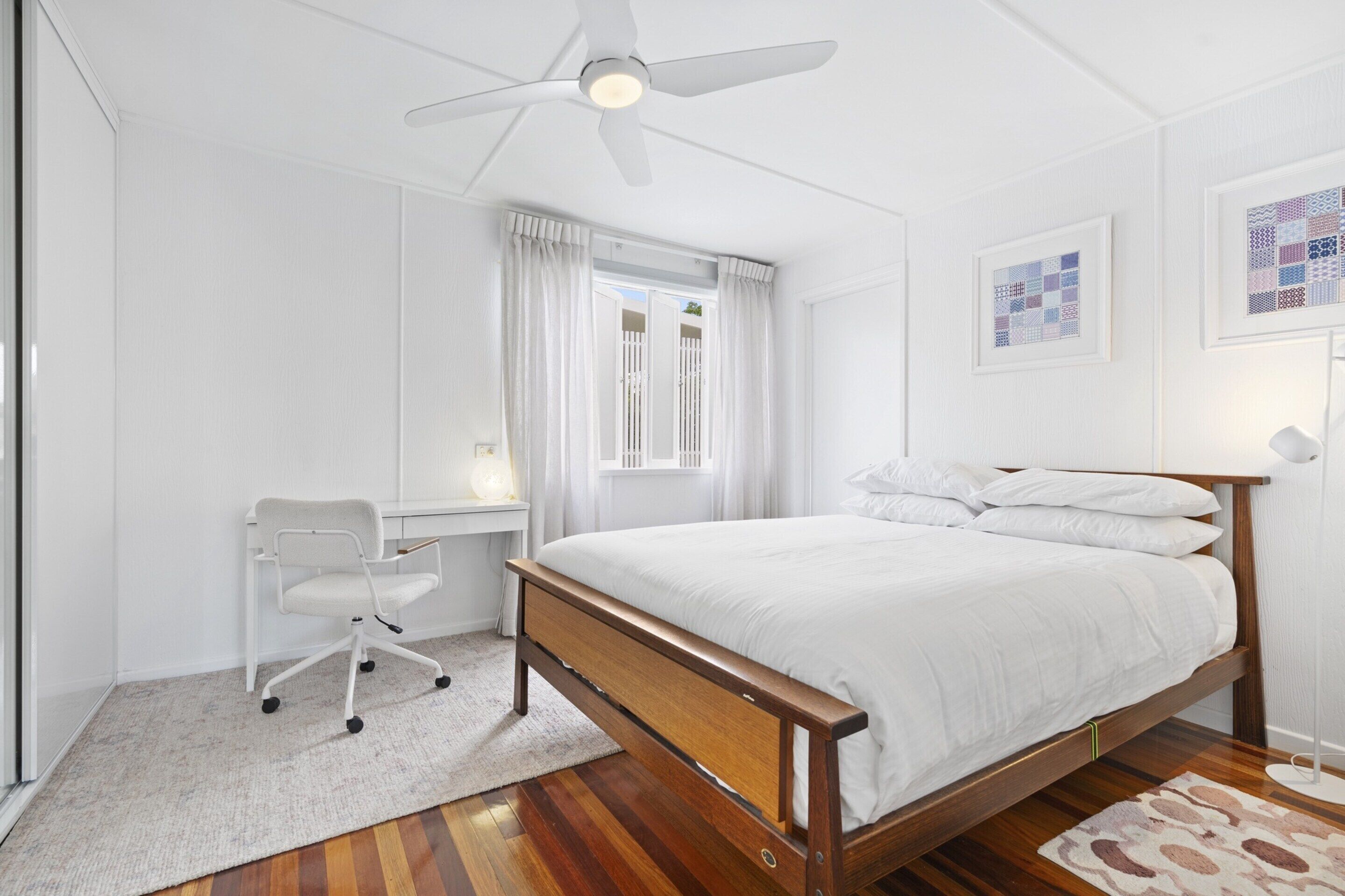Une chambre à coucher avec un lit en bois, des rideaux blancs, un bureau avec une chaise et un ventilateur de plafond.