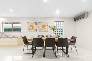 A modern dining area with a long table, black chairs, and a vase of flowers. There are two large flower paintings on the wall and an air conditioning unit above the table. - Modern 3-bed With Pool & Water Views by Beach (Sydney)
