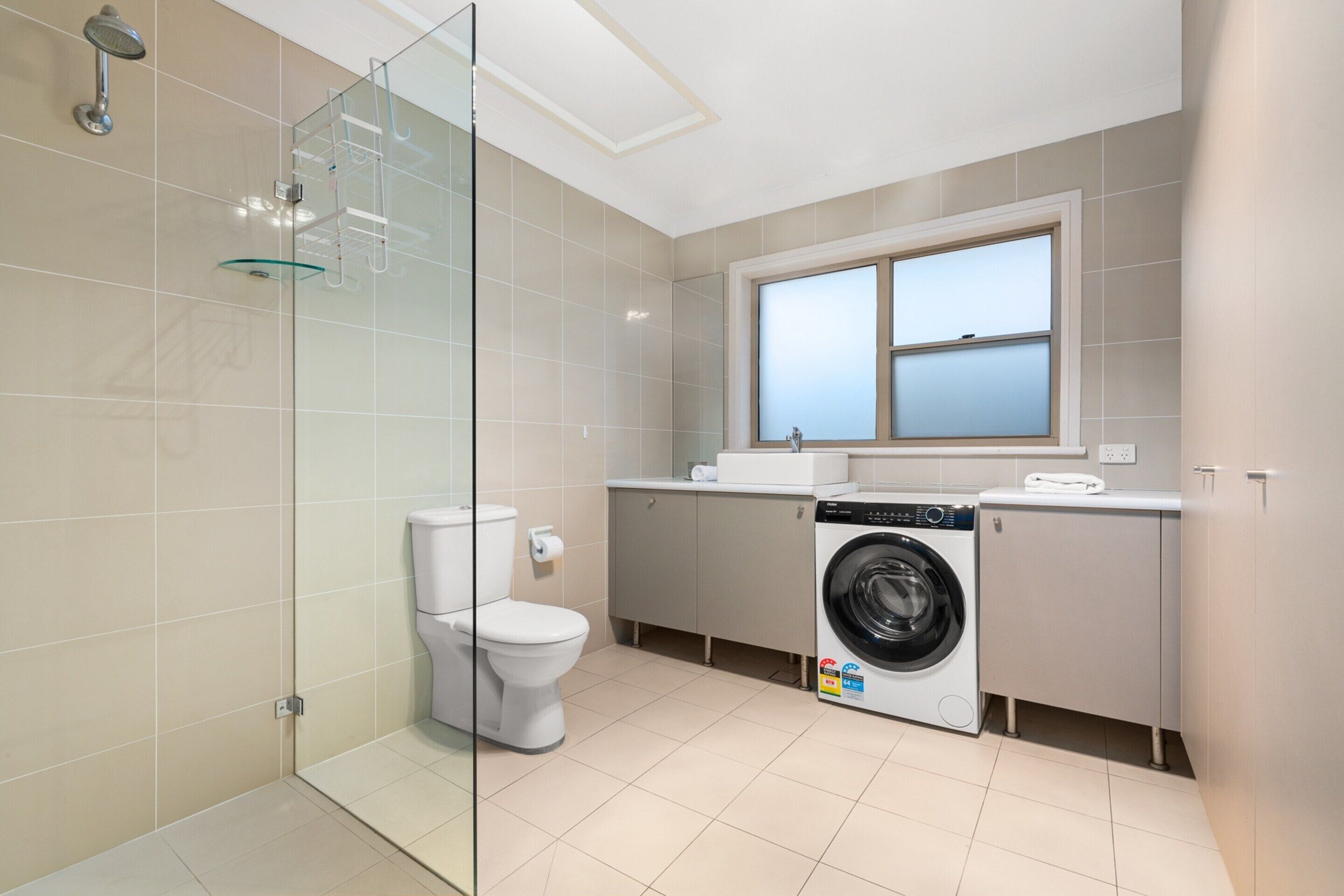 A modern bathroom with a glass shower enclosure, a toilet, a washing machine, and a window providing natural light.
