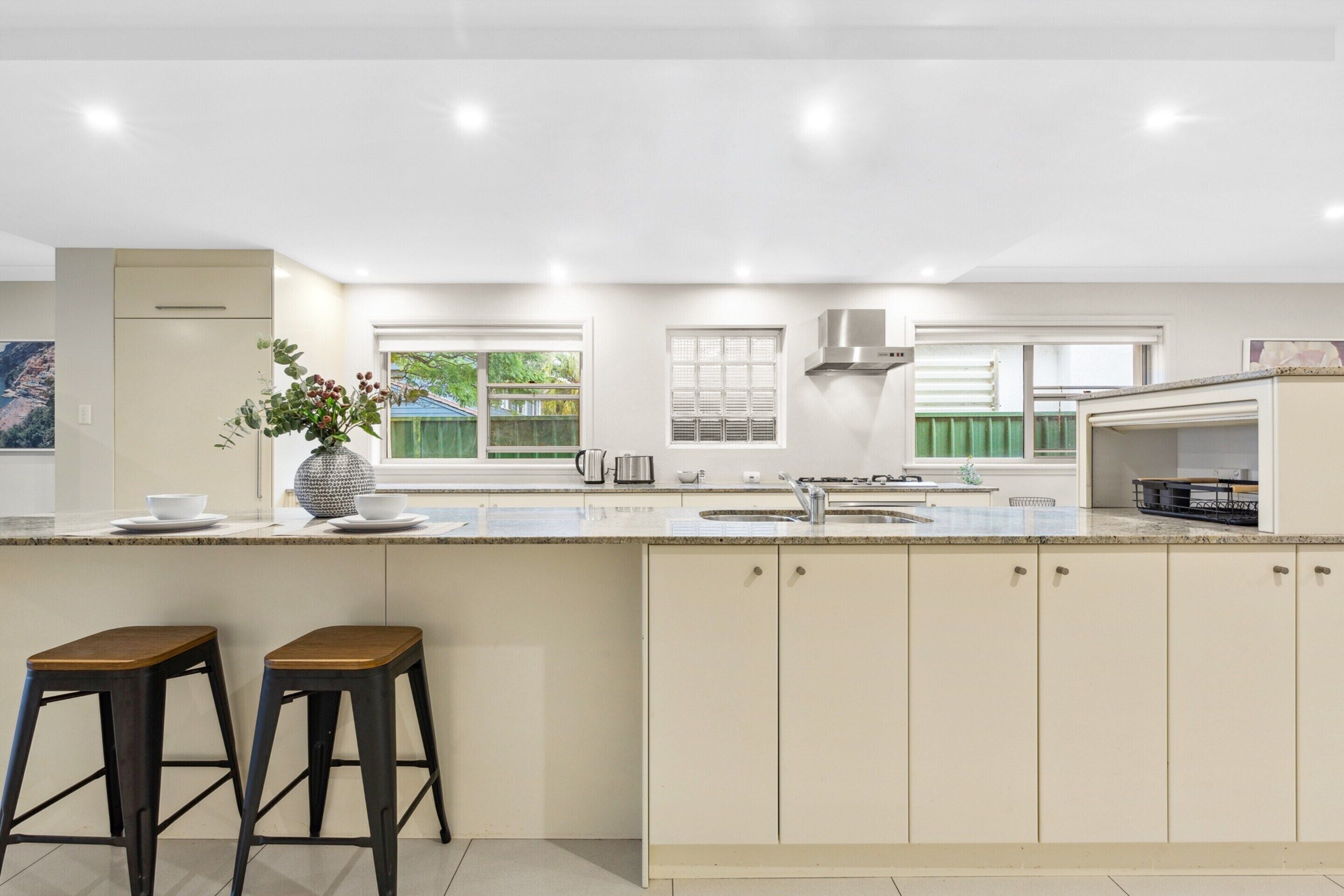 A modern kitchen with a large island, two bar stools, a built-in oven, and a window with a view of greenery.