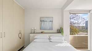 Ein Schlafzimmer mit einem Bett, einem Nachttisch, einem Gemälde und Blick auf einen Balkon mit Bäumen.