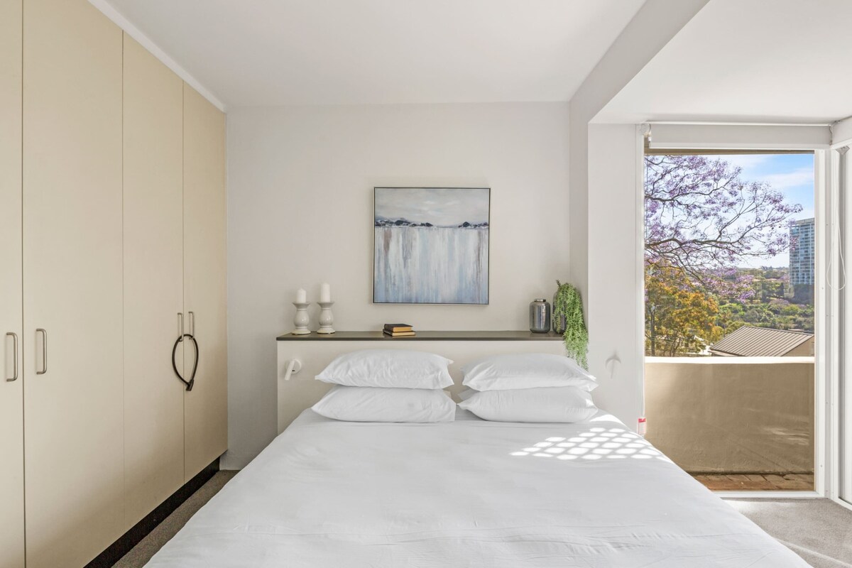 A modern bedroom with a bed, a nightstand, a painting, and a view of the outdoors.