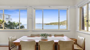 Ein Esszimmer mit großem Tisch und sechs Stühlen, großen Fenstern mit Blick auf das Meer und Hügel, sowie einer Pflanzengruppe als Mittelstück.