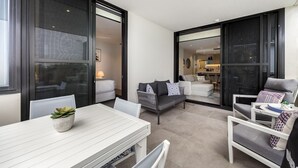 A modern outdoor patio area with a white table, chairs, and a potted plant. There is a grey sofa with patterned cushions and a white armchair with a cushion.