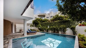 A modern outdoor swimming pool with a geometric design, surrounded by a landscaped garden and a white building with a balcony.