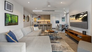 A modern living room with a sofa, coffee table, and a TV. There is a kitchen area in the background with a bar and stools.