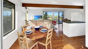 Une cuisine moderne avec une table à manger en bois, des armoires blanches et une vue sur l’océan grâce à de grandes fenêtres.