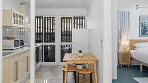 A compact room with a kitchenette, a dining area with stools, a bed with a nightstand, and a window with curtains.
