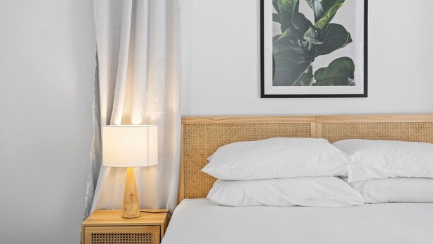 A neatly made bed with white linens, a wooden nightstand with a lamp, and a framed plant on the wall.