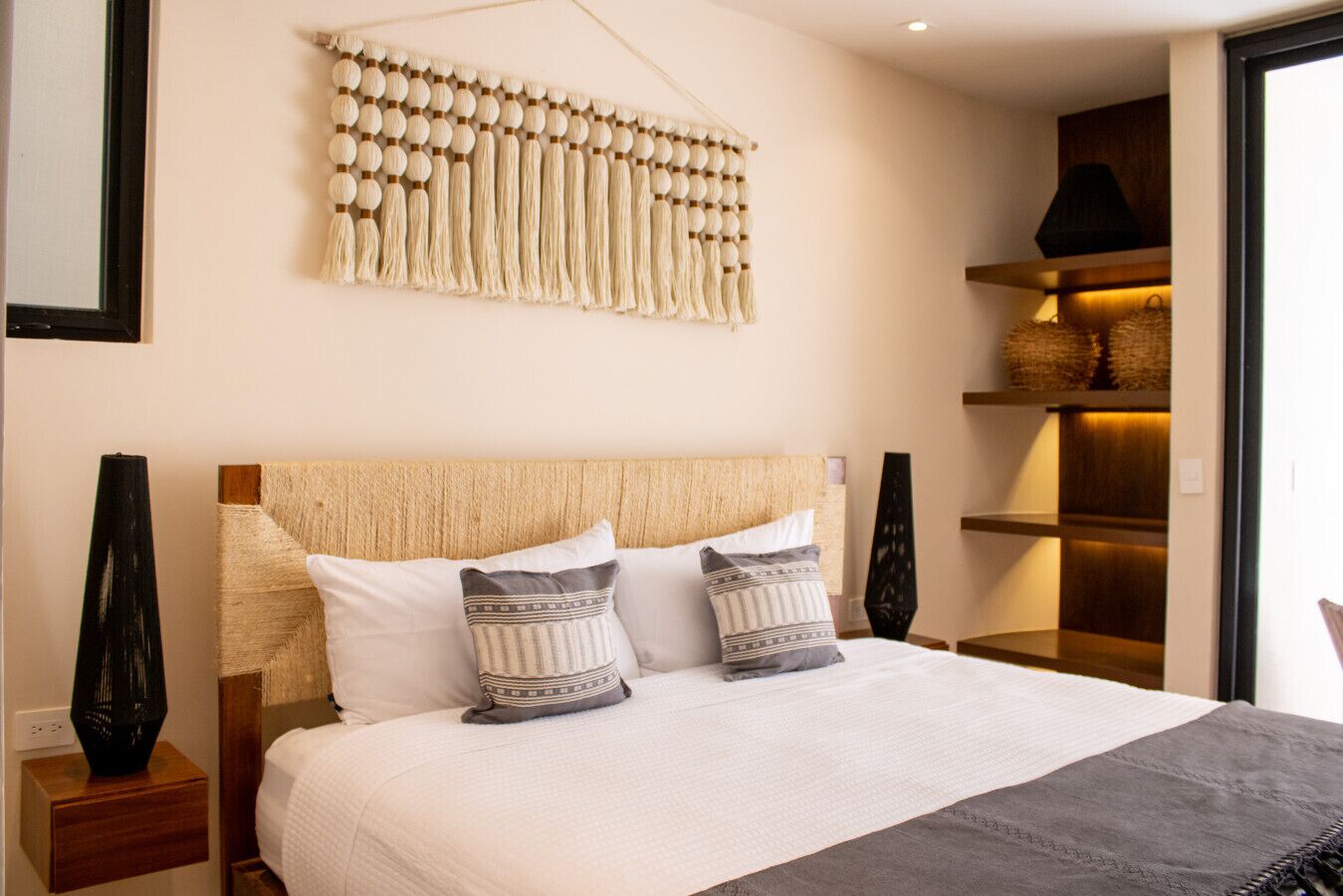 Un dormitorio con una cama grande, dos almohadas, un tapiz de macramé y una mesita de noche con un florero.