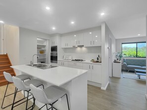 A modern kitchen with white cabinetry, a central island with bar stools, and a view of a green landscape through a large window. - Spacious 4-bed House Near Batemans Bay Centre (Batemans Bay)