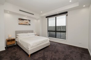A bedroom with a large bed, a nightstand, a wall-mounted air vent, a window with blinds, and a framed picture on the wall.