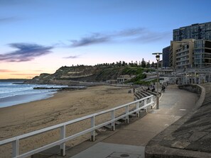 A coastal promenade with a sandy beach, a white railing, and a modern building with multiple floors. - Charming Townhouse Steps From Newcastle Beach (Newcastle)