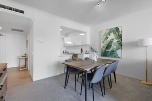 A modern kitchen with white cabinets, a dining table with chairs, and a large botanical print on the wall. - Ocean View 2-bed With Pool, Tennis Court & BBQ (Gold Coast)