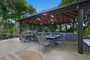 An outdoor patio with a covered dining area, surrounded by palm trees and greenery. - Ocean View 2-bed With Pool, Tennis Court & BBQ (Gold Coast)