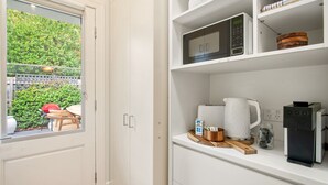 Eine Küche mit weißen Schränken, einer Mikrowelle, einer Kaffeemaschine und einem Fenster mit Blick auf Grünflächen und einem roten Stuhl draußen.