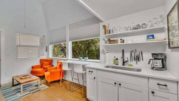 Een moderne keuken met witte kastjes, een spoelbak en diverse keukengerei. Er is een raam met zonwering, een stoel en een tafel met een boek.