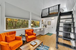 Un espacio habitable compacto con kitchenette, una zona de comedor con sillas naranjas, una mesa de centro de madera y una escalera de metal negro.