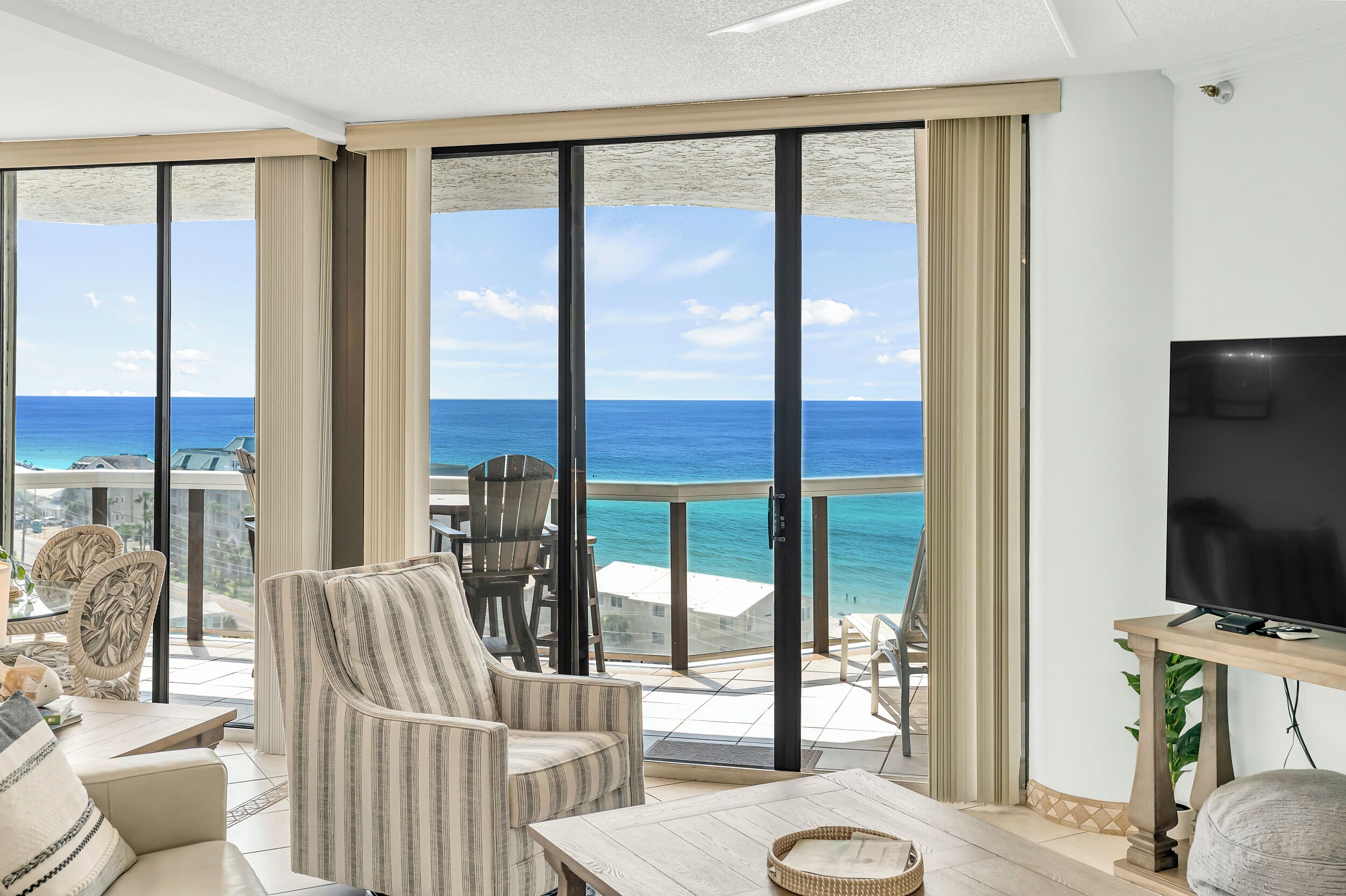 A room with a large TV, a dining table, chairs, and a view of the ocean.