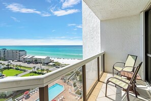 A balcony with a view of a beach and ocean, a pool, and a chair.