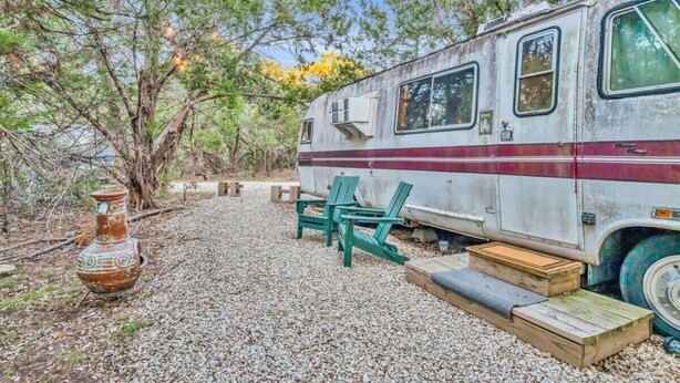 A vintage camper van parked in a natural setting with a rustic table and chairs, surrounded by trees and gravel.