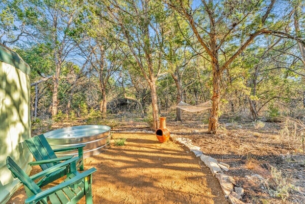 Un entorno rústico al aire libre con una hamaca, un recipiente metálico y una silla verde.