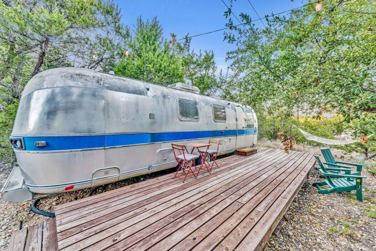 A stationary RV with a blue stripe parked on a wooden deck, surrounded by trees.