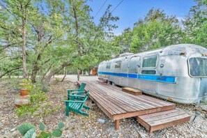 Ein alter Wohnmobilwagen steht in einem Waldgebiet mit einer Holzterrasse und grünen Stühlen.