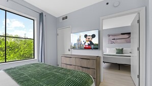 Una habitació d'hotel moderna amb un llit gran, una televisió que mostra en Mickey Mouse, vista a la vegetació a través d'una finestra i una zona d'estar acollidora amb una panxa i coixins.