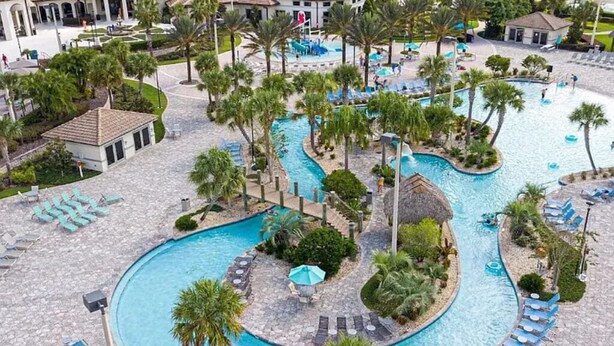 A large, winding pool surrounded by palm trees and lounge chairs, with a small building and a covered area nearby. - New 5BR/5BA home w/ private pool & hot tub, minutes to DIsney.  Sleeps up to 15. (Kissimmee)