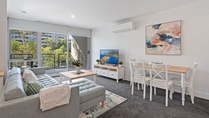 A modern living room with a grey sofa, a wooden dining table with white chairs, a flat-screen TV, and a large abstract painting.