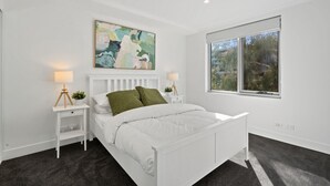 A modern bedroom with a white bed, a large abstract painting, a window with a view of trees, and a small bedside table with a lamp.