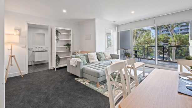 A modern living room with a grey sofa, white chairs, a dining table, and a kitchen area with white cabinets.