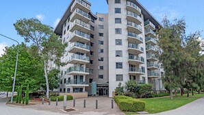 A multi-story residential building with balconies, a central entrance, and surrounding greenery.