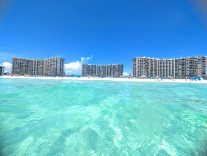 Ett hotell vid stranden med flera höga byggnader, en sandstrand och kristallklart turkost vatten.