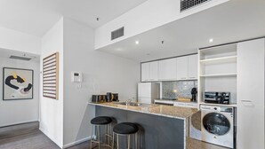 A modern kitchen with a granite countertop, white cabinets, a built-in washing machine, and two bar stools. - Chic Inner-city Stay With Balcony (Melbourne)