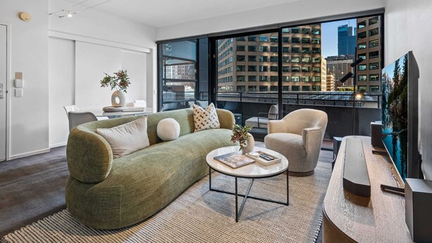 A modern living room with a green sofa, a round coffee table, and a view of tall buildings through large windows. - Chic Inner-city Stay With Balcony (Melbourne)