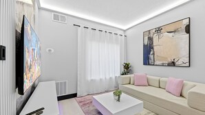 A modern living room with a flat-screen TV, a white coffee table, a beige sofa, and a large abstract painting. - 1BR Apt, 2 Queens + Sofa, Near Disney (Orlando)