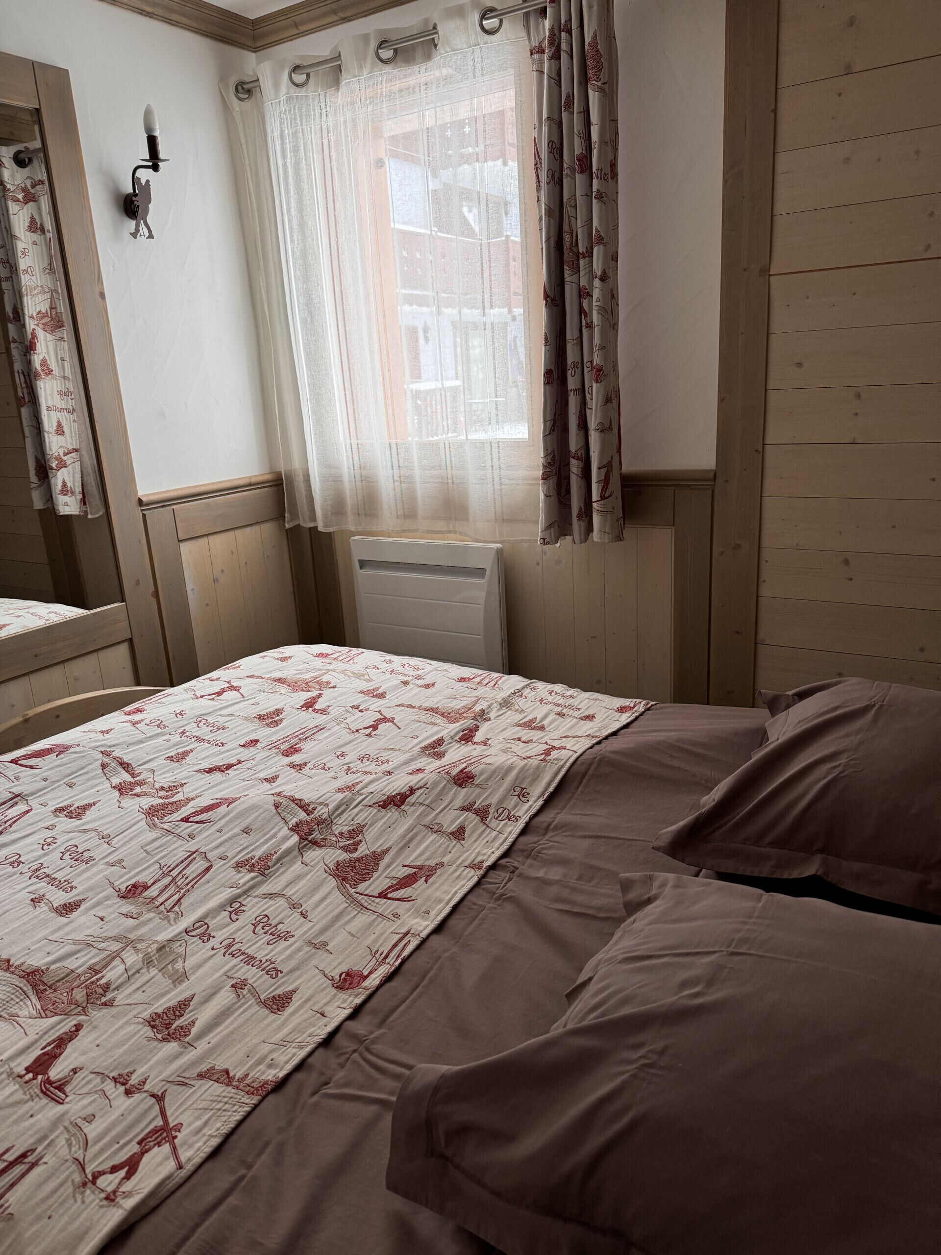 A bedroom with a bed, a window with curtains, a radiator, and a wooden wall.